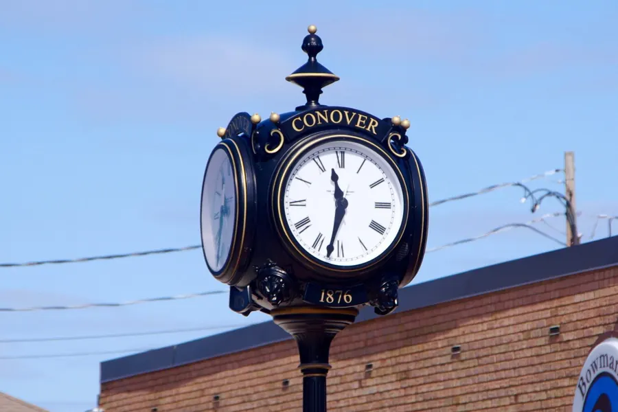 Black and gold four-sided street clock in Conover with the city name on top and the year 1876 below the face, in front of a brick storefront